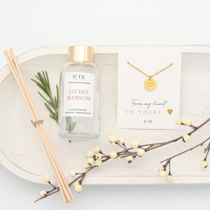May include: A white wooden tray holds a clear glass diffuser bottle with a gold cap, reeds, a sprig of rosemary, and a gold necklace with a heart pendant. The diffuser is labeled "Lychee Blossom" and the necklace card reads "From my heart to yours."