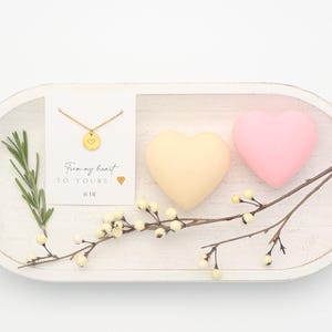 May include: A gold necklace with a heart pendant is presented on a white card with the text "From my heart to yours." Two heart-shaped objects, one yellow and one pink, are next to a sprig of rosemary and a branch with small white flowers.