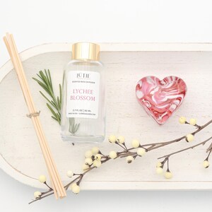May include: A clear glass bottle with a gold cap, labelled "Lychee Blossom," sits on a white wooden tray with a heart-shaped decorative piece. Reed diffuser sticks and a sprig of rosemary are also present, along with a decorative branch.