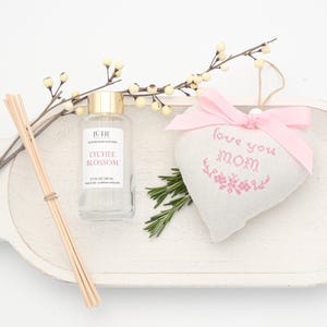 May include: A white wooden tray holds a glass bottle of Lychee Blossom diffuser, reed sticks, a heart-shaped ornament with a pink bow, and sprigs of rosemary. The ornament reads "love you mom" in pink embroidery.