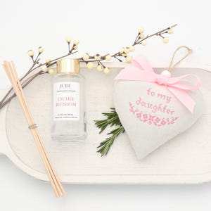 May include: A clear glass bottle with a gold cap labelled "Lychee Blossom" sits beside a heart-shaped sachet with a pink bow and the text "to my daughter". A bundle of wooden diffuser sticks and sprigs of greenery are also present.