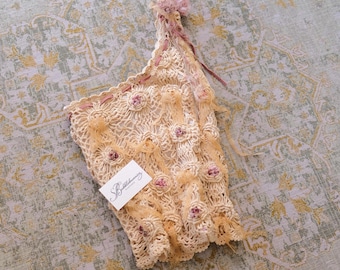 May include: An off-white, one-shoulder crochet top featuring intricate openwork patterns, small pink floral accents, and thin ribbon detailing. A white rectangular tag with the black text Bubblebunnies Vintage is attached to the front.