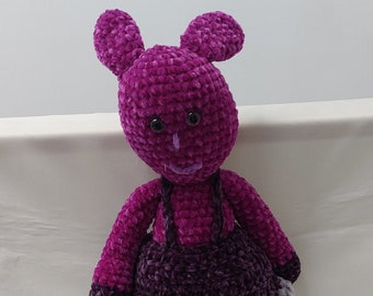 May include: A crocheted plush toy in the shape of a rabbit. The rabbit is wearing a purple dress with a gray trim and has purple and black feet. The rabbit has black eyes and a purple nose.