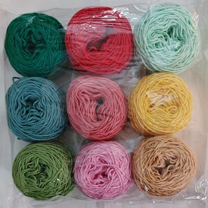 May include: Nine balls of yarn in different colors, including green, red, pink, yellow, and blue. The yarn is arranged in a circle on a white surface.