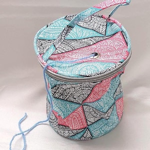 May include: A teal, pink, and black patterned yarn bowl with a handle. The bowl has a zipper closure and is made of fabric.