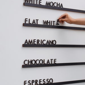 K&ouml;nnte beinhalten: Ein Wandmen&uuml;board mit f&uuml;nf horizontalen schwarzen Schienen und dunkelbraunen ausgeschnittenen Buchstaben, die WHITE MOCHA, FLAT WHITE, AMERICANO, CHOCOLATE und ESPRESSO bilden. Die Buchstaben sind einzeln auf den Schienen platziert.