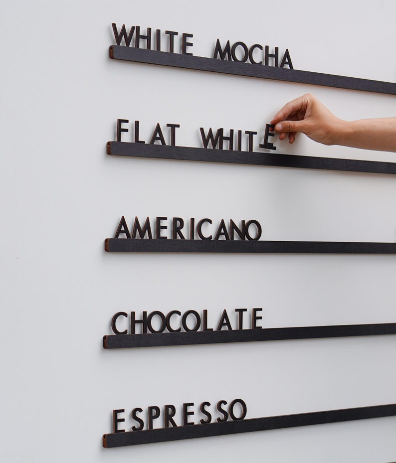 K&ouml;nnte beinhalten: Ein Wandmen&uuml;board mit f&uuml;nf horizontalen schwarzen Schienen und dunkelbraunen ausgeschnittenen Buchstaben, die WHITE MOCHA, FLAT WHITE, AMERICANO, CHOCOLATE und ESPRESSO bilden. Die Buchstaben sind einzeln auf den Schienen platziert.