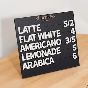 May include: A black square menu board with horizontal slots featuring white raised letters and numbers. The text reads: churrasko GRILL HOUSE, LATTE 5/2, FLAT WHITE 4, AMERICANO 3/5, LEMONADE 5, and ARABICA 6. The board has a matte finish.