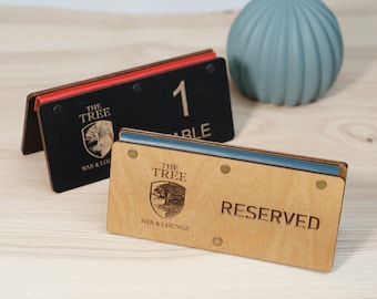 May include: Wooden table signs with the text "THE TREE BAR & LOUNGE" and a tree logo. One sign is black with the number "1" and the word "TABLE". The other sign is natural wood with the word "RESERVED".