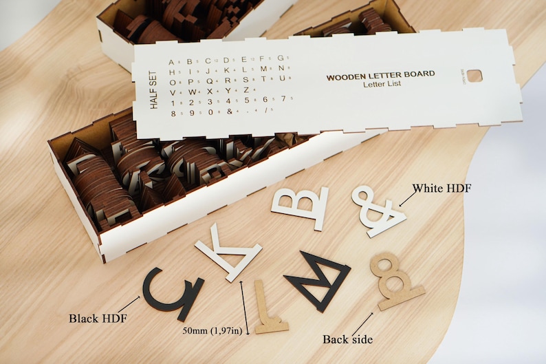 K&ouml;nnte beinhalten: Ein Holzbuchstaben-Set mit einer wei&szlig;en F&uuml;hrungskarte, die eine Alphabet- und Zahlenliste enth&auml;lt. Das Set umfasst verschiedene Buchstaben und Symbole in Schwarz, Wei&szlig; und Naturholz, aufbewahrt in einer wei&szlig;en Box. Ein Etikett zeigt 50mm an.