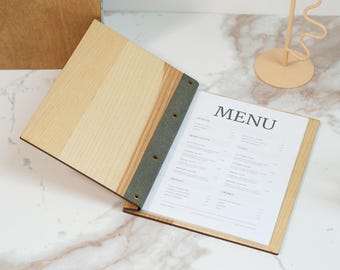 May include: Wooden menu holder with a light wood grain finish, open to display a printed menu. The menu is visible with the word "MENU" at the top. The holder is bound with a gray fabric spine and sits on a marble surface.