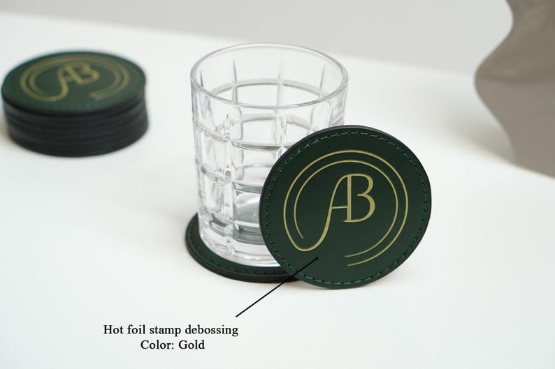 May include: Green leather coasters with a gold foil stamped debossed "AB" logo. The coasters are stacked on top of each other.