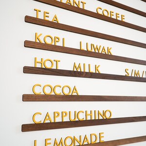 Könnte beinhalten: Eine Wandtafel mit dunklen Holzregalen und gelben Buchstaben. Das Menü listet verschiedene Getränke auf, darunter "Saft", "Instantkaffee", "Tee", "Kopi Luwak", "Heiße Milch", "Kakao", "Cappuccino" und "Limonade".