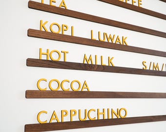May include: A wall-mounted menu board featuring horizontal dark wood rails and individual gold-colored letters. The text displays beverage options including JUICE, INSTANT COFFEE, TEA, KOPI LUWAK, HOT MILK, S/M/L, COCOA, CAPPUCHINO, and LEMONADE in a clean font.