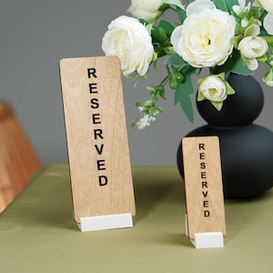 May include: Two wooden "Reserved" signs with white bases stand on a table. The signs are rectangular with the word "Reserved" printed vertically in black. A black vase with white flowers sits behind the signs.