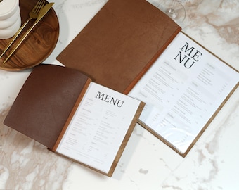May include: Brown leather menu holders with the word "MENU" printed on the white pages. The menus are open, displaying the menu items. A wooden plate with gold utensils and a white ceramic container are in the background.