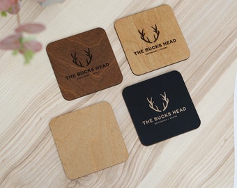 May include: Four square wooden coasters with the text "THE BUCKS HEAD RESTAURANT + ROOMS" and a deer antler logo. The coasters are in various shades of brown and black, with a light wood grain background.