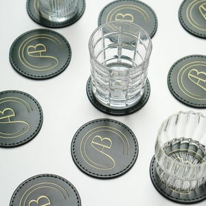 May include: Eight round green leather coasters with a gold letter "B" in a circle design. Two clear glass tumblers are on top of the coasters.