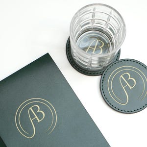 May include: Two green leather coasters with gold lettering that says "AB" and a clear glass with a gold "AB" inside. A green leather journal with gold "AB" lettering is also in the image.