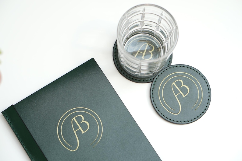 May include: Two green leather coasters with gold lettering that says "AB" and a clear glass with a gold "AB" inside. A green leather journal with gold "AB" lettering is also in the image.