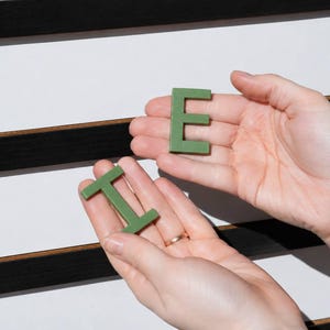 K&ouml;nnte beinhalten: Zwei H&auml;nde halten salbeigr&uuml;ne Kunststoffbuchstaben E und I vor einem wei&szlig;en Buchstabenbrett mit horizontalen schwarzen Filzrillen. Das Buchstabenbrett zeigt weitere gr&uuml;ne Buchstaben auf der Oberfl&auml;che, darunter ein teilweises E links.