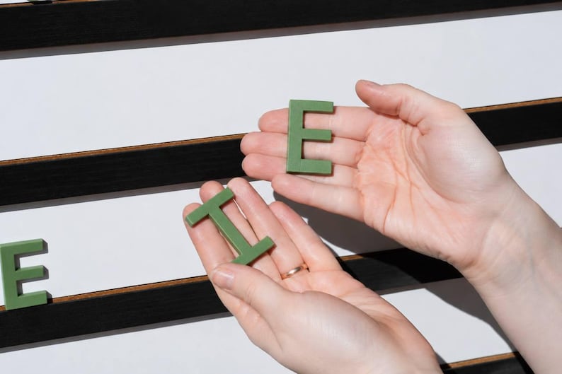 K&ouml;nnte beinhalten: Zwei H&auml;nde halten salbeigr&uuml;ne Kunststoffbuchstaben E und I vor einem wei&szlig;en Buchstabenbrett mit horizontalen schwarzen Filzrillen. Das Buchstabenbrett zeigt weitere gr&uuml;ne Buchstaben auf der Oberfl&auml;che, darunter ein teilweises E links.