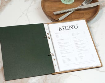 May include: Open menu binder with the word "MENU" in a black font. The menu is displayed on a white paper with a wooden frame. The left side of the binder is a dark green color. A wooden plate with silverware is in the background.