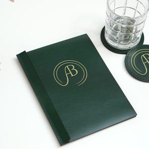 May include: A dark green leather-bound journal with a gold embossed monogram "AB" on the front cover. The journal is sitting on a white surface with two matching green leather coasters.