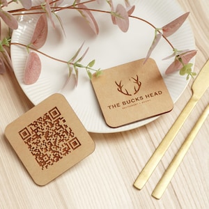 May include: Two square wooden coasters. One coaster features a QR code and the other has a logo with the text "The Bucks Head Restaurant & Rooms".
