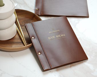 May include: A brown leather bar menu with gold lettering that reads "BELLAMONTE BAR MENU". The menu is open, revealing the inside pages. A wooden tray with a white vase and gold chopsticks is in the background.