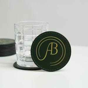 May include: A green leather coaster with a gold "AB" monogram in a circle. The coaster is sitting on a white surface next to a clear glass with a grid pattern.