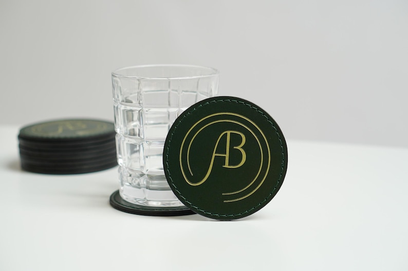 May include: A green leather coaster with a gold "AB" monogram in a circle. The coaster is sitting on a white surface next to a clear glass with a grid pattern.