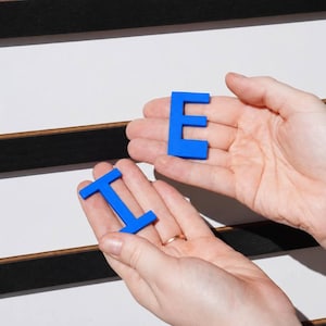 K&ouml;nnte beinhalten: Zwei leuchtend blaue Kunststoffbuchstaben, ein E und ein I, werden vor einer wei&szlig;en Tafel mit horizontalen schwarzen Filzstreifen gehalten. Die Buchstaben sind massiv, flach und in Gro&szlig;buchstaben in einer serifenlosen Schriftart gehalten.