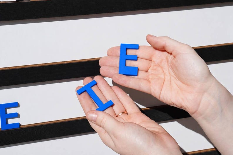 K&ouml;nnte beinhalten: Zwei leuchtend blaue Kunststoffbuchstaben, ein E und ein I, werden vor einer wei&szlig;en Tafel mit horizontalen schwarzen Filzstreifen gehalten. Die Buchstaben sind massiv, flach und in Gro&szlig;buchstaben in einer serifenlosen Schriftart gehalten.