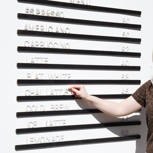 May include: A wall-mounted menu board featuring horizontal black rails with white, three-dimensional letters and numbers. The text displays items including Espresso, Americano, Cappuccino, Latte, Flat White, Chai Latte, Cold Brew, Ice Latte, and Lemonade.