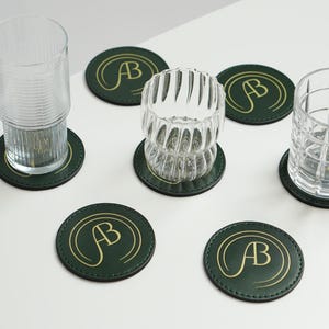 May include: Six green leather coasters with gold lettering that says "AB" in a circle. The coasters are on a white surface with two clear glasses on top of them.