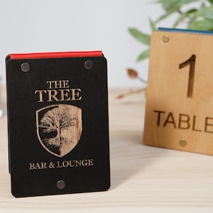 May include: Wooden table number and bar sign. The table number is light brown with the number 1 and the word TABLE engraved. The bar sign is black with the words "THE TREE BAR & LOUNGE" and a tree logo.
