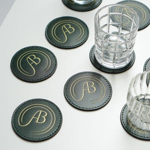 Op de afbeelding: Acht ronde groene leren onderzetters met gouden letters die "AB" zeggen op elke onderzetter. Twee heldere glazen tumblers staan op twee van de onderzetters.