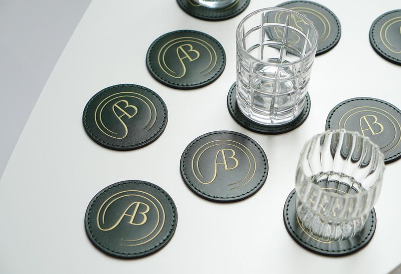 May include: Eight round green leather coasters with gold lettering that says "AB" on each coaster. Two clear glass tumblers are sitting on top of two of the coasters.