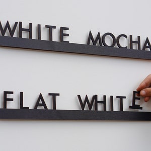 K&ouml;nnte beinhalten: Zwei horizontale schwarze Holzschienen mit einzelnen schwarzen Holzbuchstaben. Die obere Schiene zeigt den Text WHITE MOCHA. Die untere Schiene zeigt den Text FLAT WHITE, wobei der letzte Buchstabe E von einer Hand gehalten wird.
