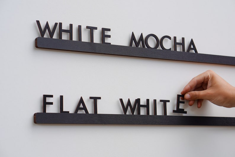 K&ouml;nnte beinhalten: Zwei horizontale schwarze Holzschienen mit einzelnen schwarzen Holzbuchstaben. Die obere Schiene zeigt den Text WHITE MOCHA. Die untere Schiene zeigt den Text FLAT WHITE, wobei der letzte Buchstabe E von einer Hand gehalten wird.