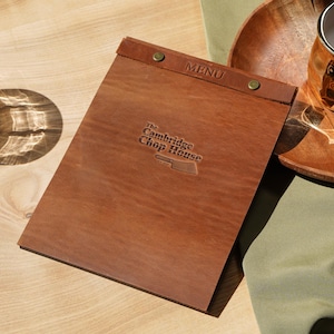 May include: A rectangular brown leather menu cover featuring a top binding strip secured with two brass-colored metal rivets. The word MENU is embossed on the top strip, and The Cambridge Chop House is embossed in the center with a cleaver icon.