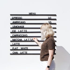 May include: A wall-mounted black menu board with horizontal rails featuring white lettering for items including Espresso, Americano, Lemonade, Ice Latte, Cappuccino, Matcha Latte, Latte, Flat White, and Chai Latte, with prices 3.0 and 3.5.