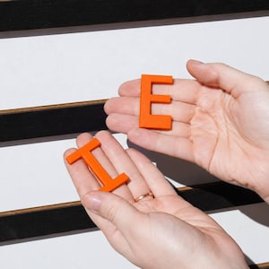 K&ouml;nnte beinhalten: Leuchtend orangefarbene Kunststoffbuchstaben E und I, die in den Handfl&auml;chen vor einer wei&szlig;en Tafel mit horizontalen schwarzen Leisten gehalten werden. Die Buchstaben sind massiv, flach und in Gro&szlig;buchstaben. Das E liegt in der oberen Handfl&auml;che, das I in der unteren, ein weiteres orangefarbenes E ist auf der Tafel zu sehen.