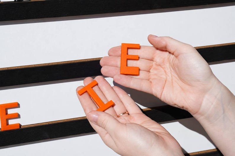 K&ouml;nnte beinhalten: Leuchtend orangefarbene Kunststoffbuchstaben E und I, die in den Handfl&auml;chen vor einer wei&szlig;en Tafel mit horizontalen schwarzen Leisten gehalten werden. Die Buchstaben sind massiv, flach und in Gro&szlig;buchstaben. Das E liegt in der oberen Handfl&auml;che, das I in der unteren, ein weiteres orangefarbenes E ist auf der Tafel zu sehen.