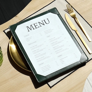 May include: A restaurant menu with the word "MENU" at the top. The menu is in a dark green leather-like cover, with a gold plate and gold utensils. A green glass is on the left side of the image.