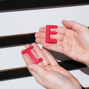 K&ouml;nnte beinhalten: Zwei leuchtend rote Kunststoffbuchstaben, ein E und ein I, werden in offenen Handfl&auml;chen gehalten. Die Buchstaben sind massiv, flach und serifenlos. Im Hintergrund befindet sich ein wei&szlig;es Steckbrett mit horizontalen schwarzen Rillen.