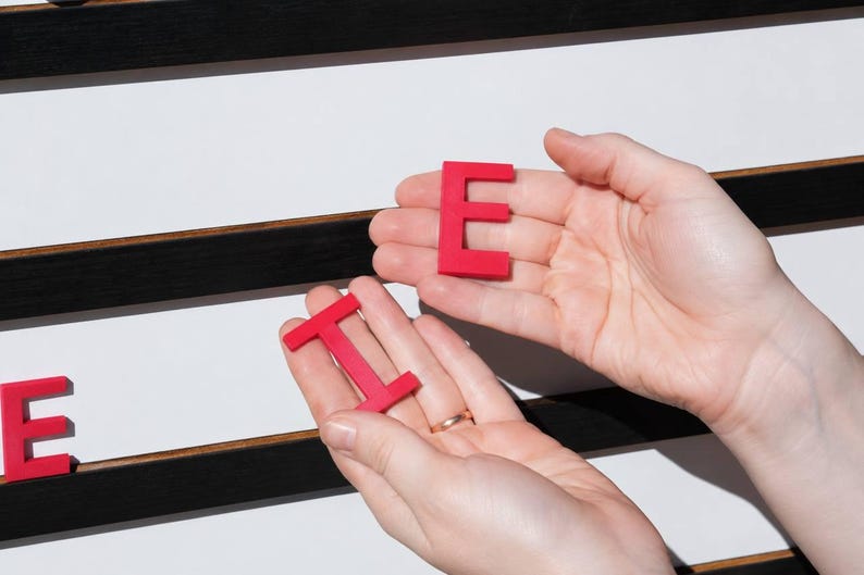 K&ouml;nnte beinhalten: Zwei leuchtend rote Kunststoffbuchstaben, ein E und ein I, werden in offenen Handfl&auml;chen gehalten. Die Buchstaben sind massiv, flach und serifenlos. Im Hintergrund befindet sich ein wei&szlig;es Steckbrett mit horizontalen schwarzen Rillen.