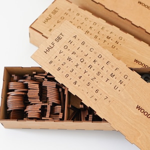 K&ouml;nnte beinhalten: Eine rechteckige Aufbewahrungsbox aus Holz mit Trennw&auml;nden, die zahlreiche kleine, dunkelbraune Holzbuchstaben und Zahlen enth&auml;lt. Ein schiebbarer Holzdeckel liegt oben auf und zeigt ein aufgedrucktes Raster mit der Beschriftung HALF SET.