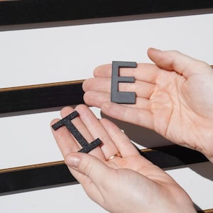 K&ouml;nnte beinhalten: Zwei schwarze, texturierte, dreidimensionale Buchstaben, ein E und ein I, in den Handfl&auml;chen gehalten. Die Buchstaben bestehen aus einem dunklen, k&ouml;rnigen Material und befinden sich vor einer wei&szlig;en Tafel mit schwarzen Lamellen.